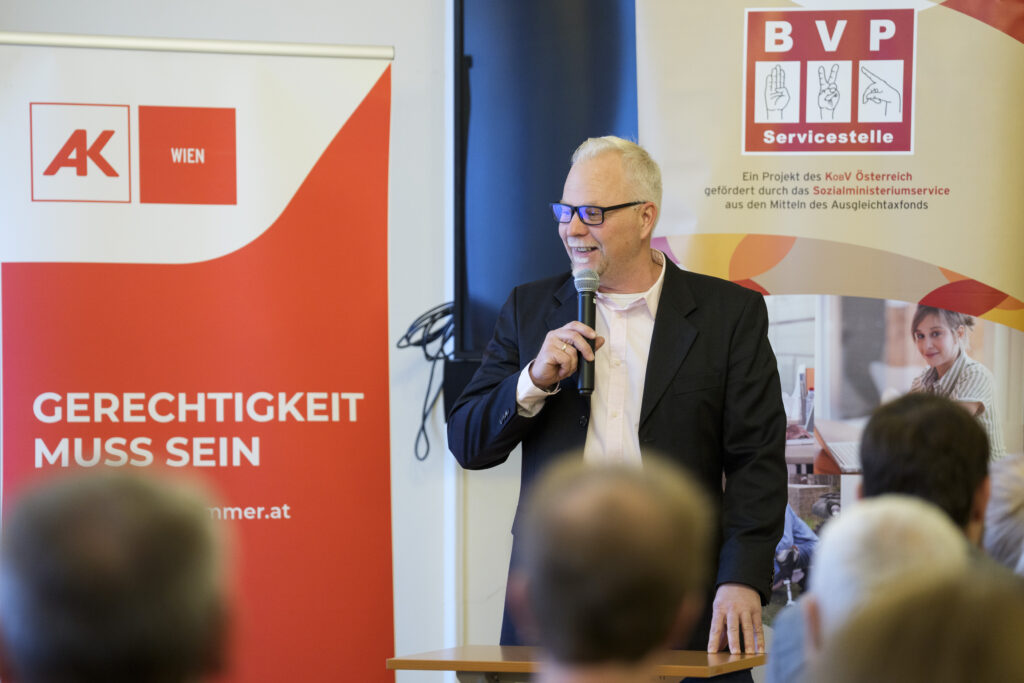 Ein Mann, Josef Bezeredj-Babarczy von der BVP-Servicestelle, spricht vor einer Gruppe Menschen in ein Mikrofon.