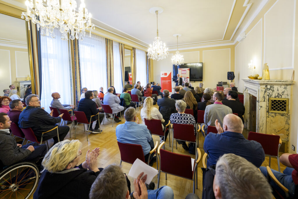 Ein Mann, KOBV-Präsident Franz Groschan, spricht vor einer Gruppe Menschen, die auf Sesseln sitzen, in ein Mikrofon.