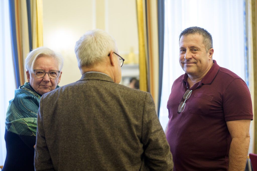 Ein Mann sieht während der Unterhaltung mit zwei anderen Menschen in die Kamera.